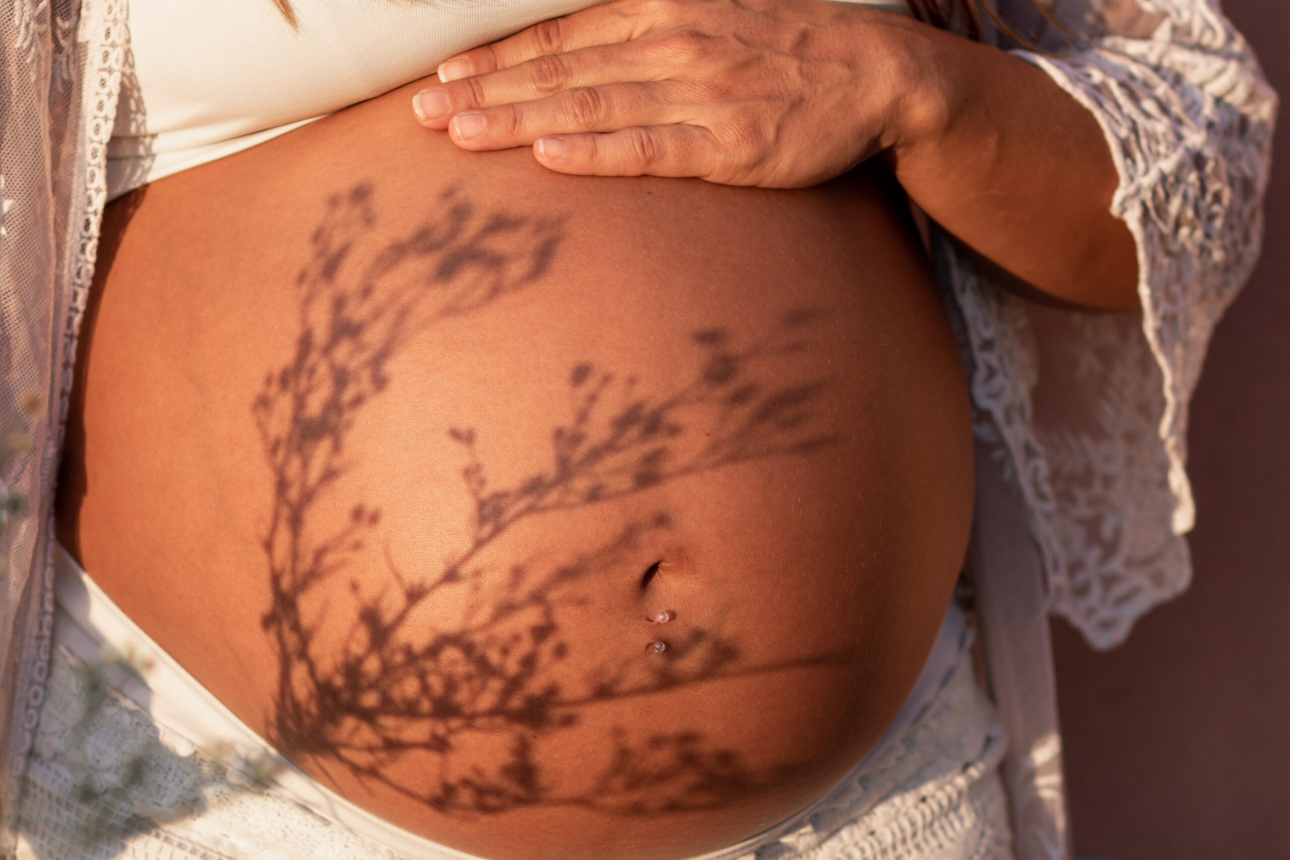 A pregnant woman with laying hand on her belly is standing against the sun and the shadow of a herbal tree soothingly covers part of the mother's belly. Herbals and natural oils used to comfort the pregnant woman.